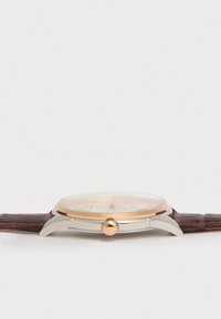 Στρογγυλό ρολόι με διαφανή πρόσωπο, ατσάλινο σώμα σε ασημί χρώμα, χρυσές λεπτομέρειες και σκούρο καφέ δερμάτινο λουράκι με λεπτομερές σχέδιο.