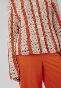 Häkeljacke mit vertikalen cremefarbenen und orangenen Streifen, ausgestattet mit großen weißen Knöpfen und einem strukturierten Strickdesign, kombiniert mit orangenen Hosen.