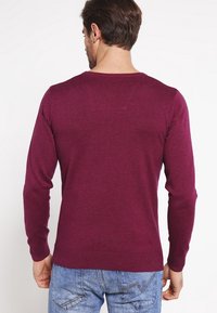 Pull en maille bordeaux à manches longues avec une encolure ronde, des poignets et un ourlet côtelés ; présente un petit détail en patch sur le haut du dos.