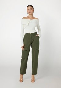 Top bianco a spalle scoperte in costina, pantaloni cargo verde oliva con tasche multiple, dettaglio con bottoni dorati e tacchi trasparenti con cinturini.