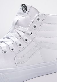 High-top white canvas sneakers with round laces, metal eyelets, and a rubber sole. Features stitched accents and a branded tag on the top.