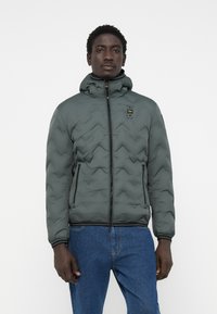 Quilted puffer jacket in dark gray with a hood, zip closure, and side pockets. Features a textured zigzag pattern and contrasting black trim.