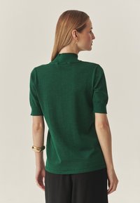 Zielona koszulka polo z krótkim rękawem o teksturowanym wykończeniu, z kołnierzem stójką i ściągaczami na mankietach. Noszona z czarnymi spodniami.