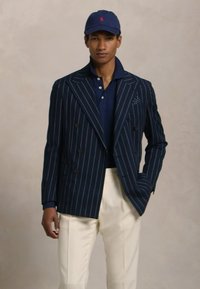 Blazer riscado azul-marinho com design de duplo peito, botões brilhantes e um lenço de bolso. Usado sobre uma polo azul-marinho e combinado com calças creme.