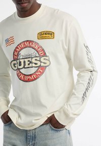 Crème lange mouwen shirt met een groot rood en zwart "GUESS" logo, gele label en accent van de Amerikaanse vlag. Katoenen stof, relaxte pasvorm.