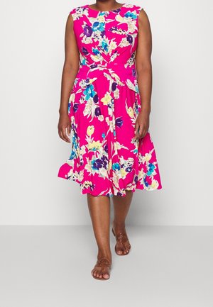 Kobieta w jasnoróżowej, bezrękawnikowej sukience z niebieskimi, żółtymi i białymi kwiatowymi wzorami, chodząca boso po gładkiej, jasnej podłodze.