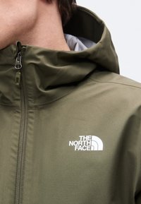 Κοντινό πλάνο ενός ατόμου που φοράει ένα χακί μπουφάν North Face με κουκούλα και μπροστινό φερμουάρ, με το λογότυπο ορατό στο αριστερό στήθος.