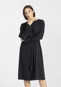 Czarna sukienka z długimi, marszczonymi rękawami, z teksturowanymi detalami. Prosty krój, pasek w talii, długość do kolan, gładki materiał, minimalistyczny design.