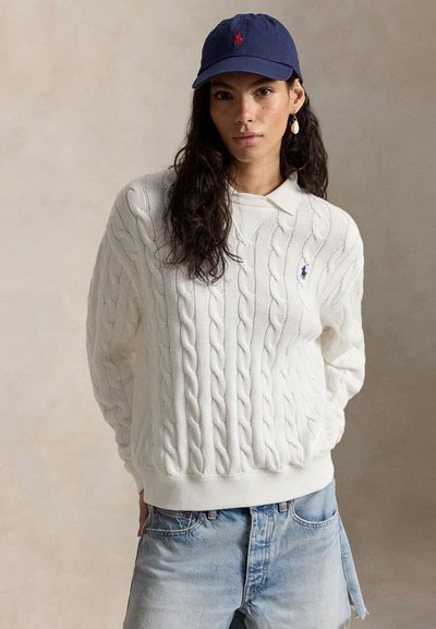 Frau mit langen dunklen Haaren, die einen weißen Strickpullover mit Zopfmuster, hellblaue Jeansshorts, eine blaue Baseballkappe und einen baumelnden Ohrring trägt, nach vorne blickend.