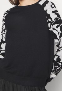 Svart stickad tröja med vita mönstrade ärmar, med rundad halsringning, ribbade manschetter och nederkant. Materialet ser mjukt och texturerat ut.