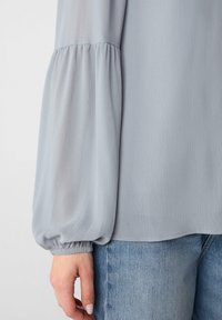 Hellblause Bluse mit puffierten Ärmeln, die eine subtile gerippte Textur und elastische Bündchen aufweist. Kombiniert mit hellblauen Jeans.