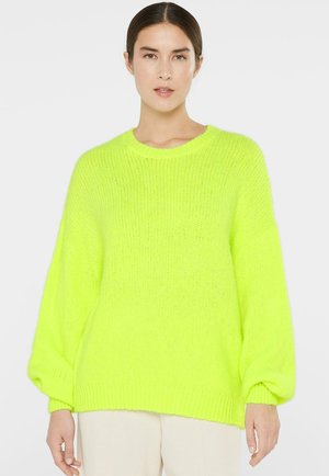 Frau trägt einen leuchtend neongelben, übergroßen Strickpullover mit langen Ballonärmeln und cremefarbenen Hosen, steht vor einem schlichten Hintergrund.