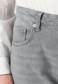 Se muestran unos vaqueros de cintura alta grises, combinados con una camisa blanca de manga larga. Un bolsillo frontal es visible, destacando los detalles de la prenda.