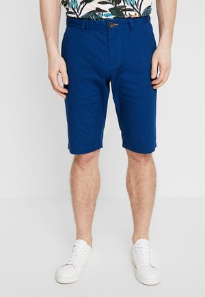 Uomo che indossa pantaloncini blu fino al ginocchio, scarpe da ginnastica bianche e una camicia con stampa tropicale, in piedi contro uno sfondo chiaro e uniforme.