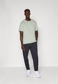 Moški stoji ob enobarvnem ozadju, oblečen v svetlo zeleno majico Nike, črne trenirka hlače Nike in bele superge.