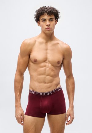 Μυώδης νέος με σγουρά μαλλιά, φορώντας μπορντό μπόξερ της Guess, στέκεται μπροστά σε ένα απλό ανοιχτό γκρι φόντο.
