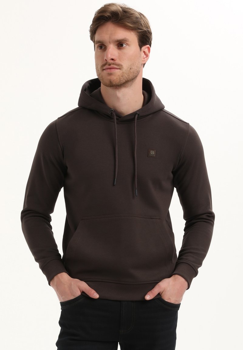 Moški v temno rjavi kapuci s sprednjim žepom in vrvicami, roke v žepih, gleda na stran pred belo ozadjem.
