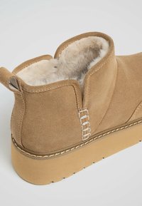 Botte cheville en daim fauve avec doublure en fourrure beige, semelle épaisse en caoutchouc et détail de couture décorative sur le côté et la languette arrière.