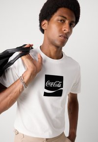 Witte t-shirt met korte mouwen, voorzien van een zwart Coca-Cola logo op een witte vierkant. Glad materiaal met ronde hals. Draagt een zwarte tas.