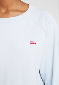 Světle modrá mikina s širokým výstřihem a raglánovými rukávy. Obsahuje malou červenou nášivku s logem Levi's na hrudi. Měkká textura látky.