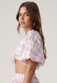 Lila rutigt kort topp med korta puffärmar, med en knytdetalj fram. Tillverkad av lätt texturerad tyg.