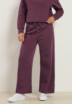 Person wearing plum-colored wide-leg sweatpants with a drawstring waist and white sneakers standing against a plain background.