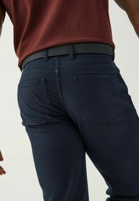 Tummansiniset trikoot, joissa on slim fit -muotoilu. Niissä on sileä pinta, kaksi takataskua ja musta vyö, joka näkyy vyötäröllä.