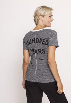 Woman with short blonde hair smiling, wearing a black and white striped T-shirt with "HUNDRED YEARS" printed on the back and black pants.