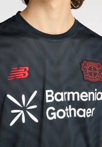 Camiseta deportiva negra con superficie texturizada, logo bordado en rojo de Bayer Leverkusen y texto blanco de Barmenia Gothaer.
