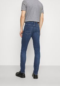 Tmavě modré úzké džíny z denimu, s klasickým designem pěti kapes a hnědou koženou nášivkou na zadním pásku.