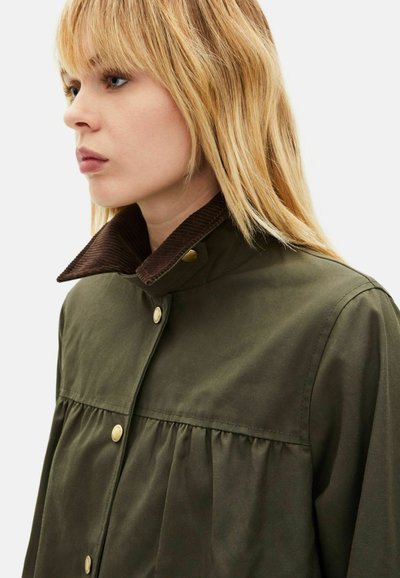 Chaqueta verde oliva con cuello de pana marrón, botones de presión y detalles plisados. Tela lisa con un ajuste estructurado, vista lateral.