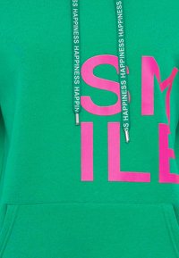 Felpa verde con grande testo rosa "SMILE". Ha una tasca frontale e un cappuccio con lacci decorati da testo ripetuto "HAPPINESS". Materiale in misto cotone.