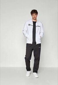En ung mann står iført en hvit jakke med glidelås, svart skjorte, svarte joggebukser med logo, og hvite joggesko mot en enkel lys bakgrunn.
