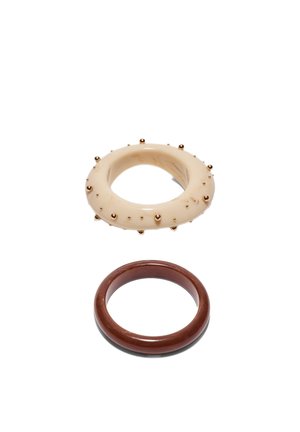 Dve okrogli zapestnici na belem ozadju; zgornja bež z zlatimi zatiči, spodnja gladka temno rjava.