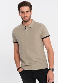 Béžové polo tričko s černým límcem a manžetami, se třemi knoflíkovými lištami a měkkou, texturovanou tkaninou, nošené s černými džíny.