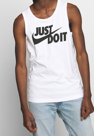 Mężczyzna w białym bezrękawniku z czarnym napisem "Just Do It" i logo Nike Swoosh, w połączeniu z jasnoniebieskimi jeansami.
