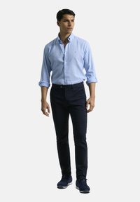 Camisa de botón azul claro, pantalones navy y zapatillas oscuras. La camisa tiene collar y mangas enrolladas, mientras que los pantalones son ajustados.