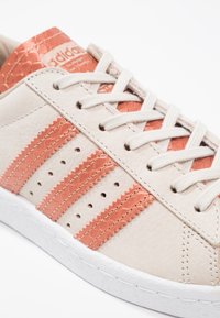 Adidas sneakers met een beige leren bovenwerk, koraalrode strepen, textuuraccenten en een witte rubberen zool. De veters zijn lichtgrijs.