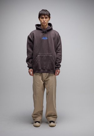 Młody mężczyzna ubrany w ciemną, za dużą bluzę z kapturem z przednią kieszenią, beżowe szerokie spodnie i beżowe trampki na jednolitym szarym tle.