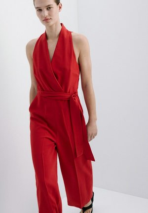 Mujer con mono rojo sin mangas de estilo envolvente, pantalones anchos y cinturón de lazo lateral, posando frente a un fondo blanco liso.