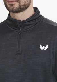 Sort lynlås sweatshirt med høj krave, struktureret stof og et hvidt logo på brystet. Lynlås og syninger er synlige.