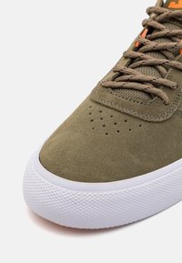 Teniska olivově zelené barvy s horní částí z broušené kůže, kulatou špičkou, perforovanými detaily a kontrastními tkaničkami. Bílá gumová podrážka s texturovaným vzorem.