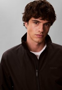 Νέος άνδρας με κυματιστά σκουρόχρωμα μαλλιά, ντυμένος με μαύρο μπουφάν με φερμουάρ πάνω από λευκό πουκάμισο, κοιτάζει ευθέως την κάμερα με φόντο ένα γκρι υπόβαθρο.