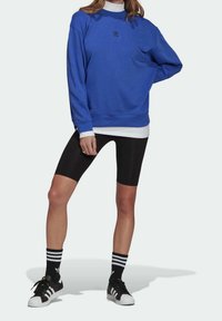 Felpa blu con polsini e orlo a costine, caratterizzata da un piccolo logo nero. Abbinata a pantaloncini da ciclismo neri e scarpe nere con dettagli bianchi.