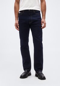 Σκούρο μπλε τζιν παντελόνι με ίσια γραμμή, με πορτοκαλί ραφές, μπροστινές τσέπες και κλείσιμο με κουμπιά. Συνδυάζεται με μαύρα παπούτσια.