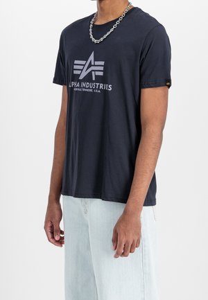 Mand iført en sort Alpha Industries T-shirt med logo og tekst, lyseblå jeans og en sølvkæde mod en hvid baggrund.