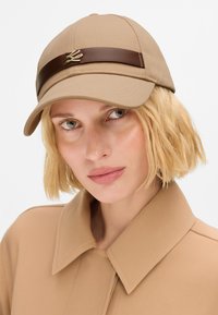 Beige baseball cap with a brown leather band and a gold accent. The cap has a structured shape and a smooth fabric texture.