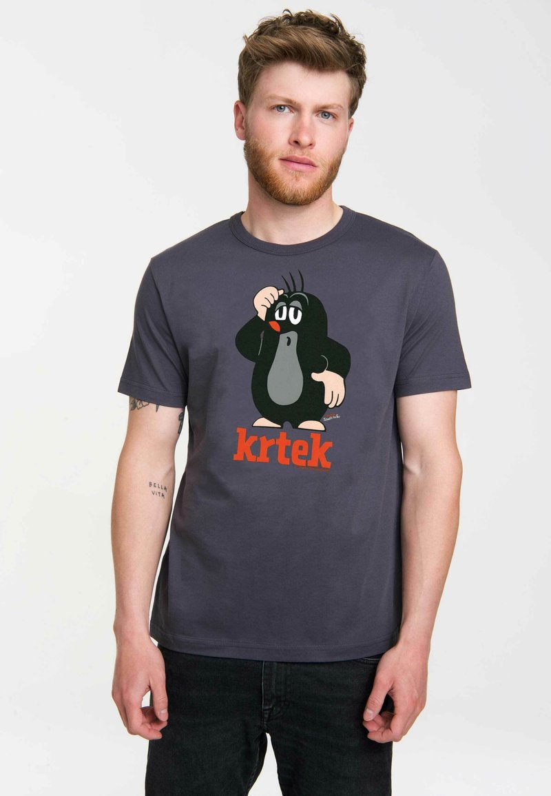 Young man with light brown hair and beard wearing dark gray t-shirt with cartoon mole character and red text "krtek" on front.