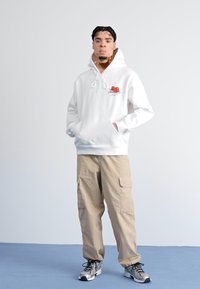 Hvit hettegenser med kirsebærgrafikk, kombinert med beige cargo-bukser. Modell står med hendene i lommene på en blå overflate.