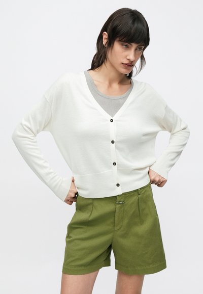 Λευκή ζακέτα με V-λαιμόκοψη, με μαύρα κουμπιά, συνδυασμένη με σορτς σε ελιά πράσινο. Η ζακέτα έχει άνετη εφαρμογή και μακριά μανίκια.